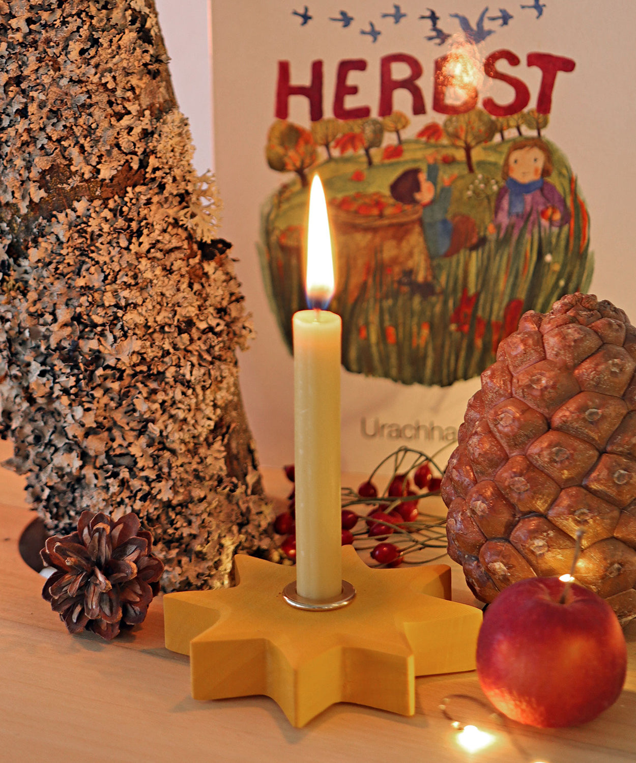 Grimm's Yellow Lifelight Star with a lit candle set up in a little seasonal display featuring a small apple and pine cones. 