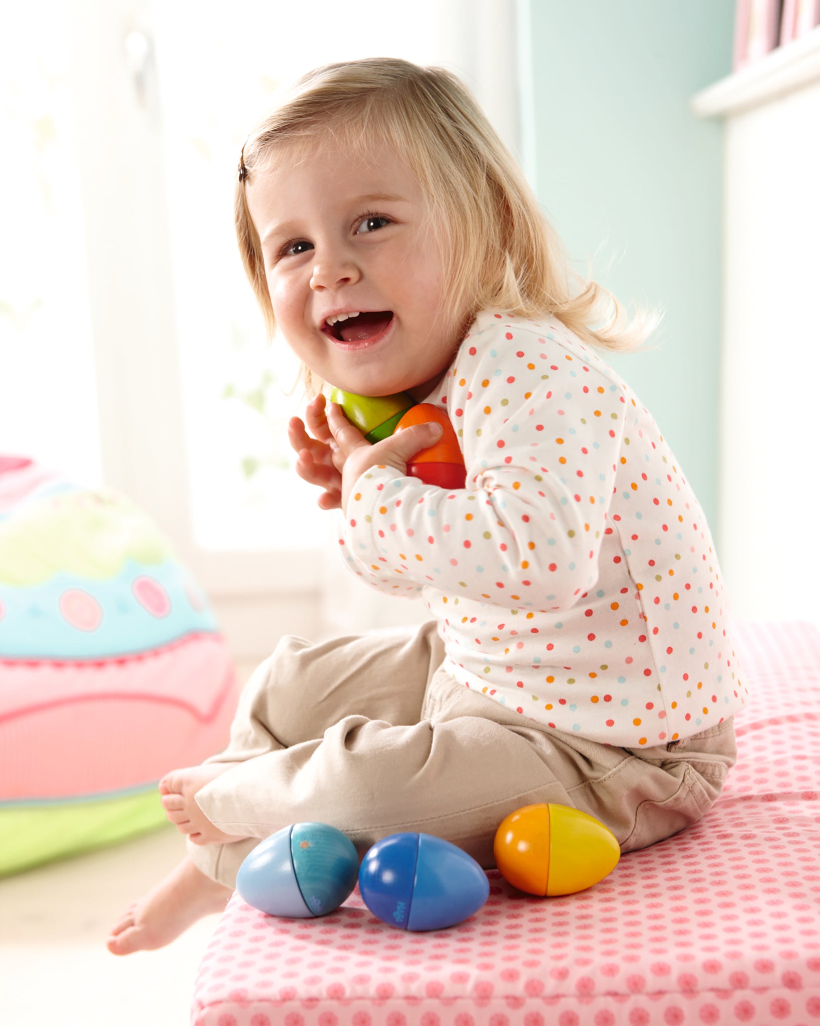 A child chewing on the blue Haba Musical Wooden Egg Toy whilst sat on a light blue rug with the other Haba Musical Eggs by their feet