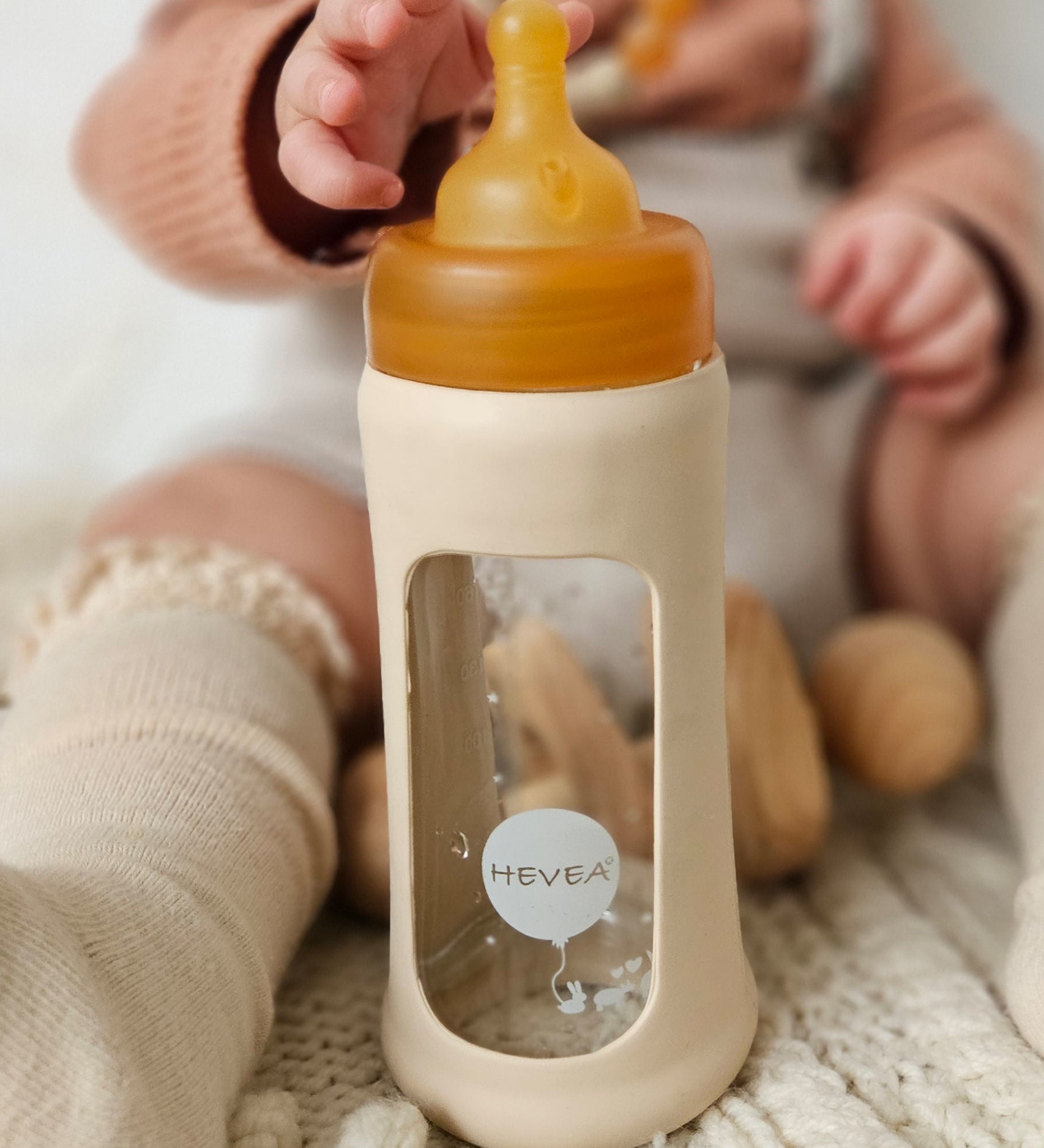 Baby sat on the floor reaching towards the Hevea Glass Bottle Wide Neck With Rubber Sleeve - 250ml in Sand