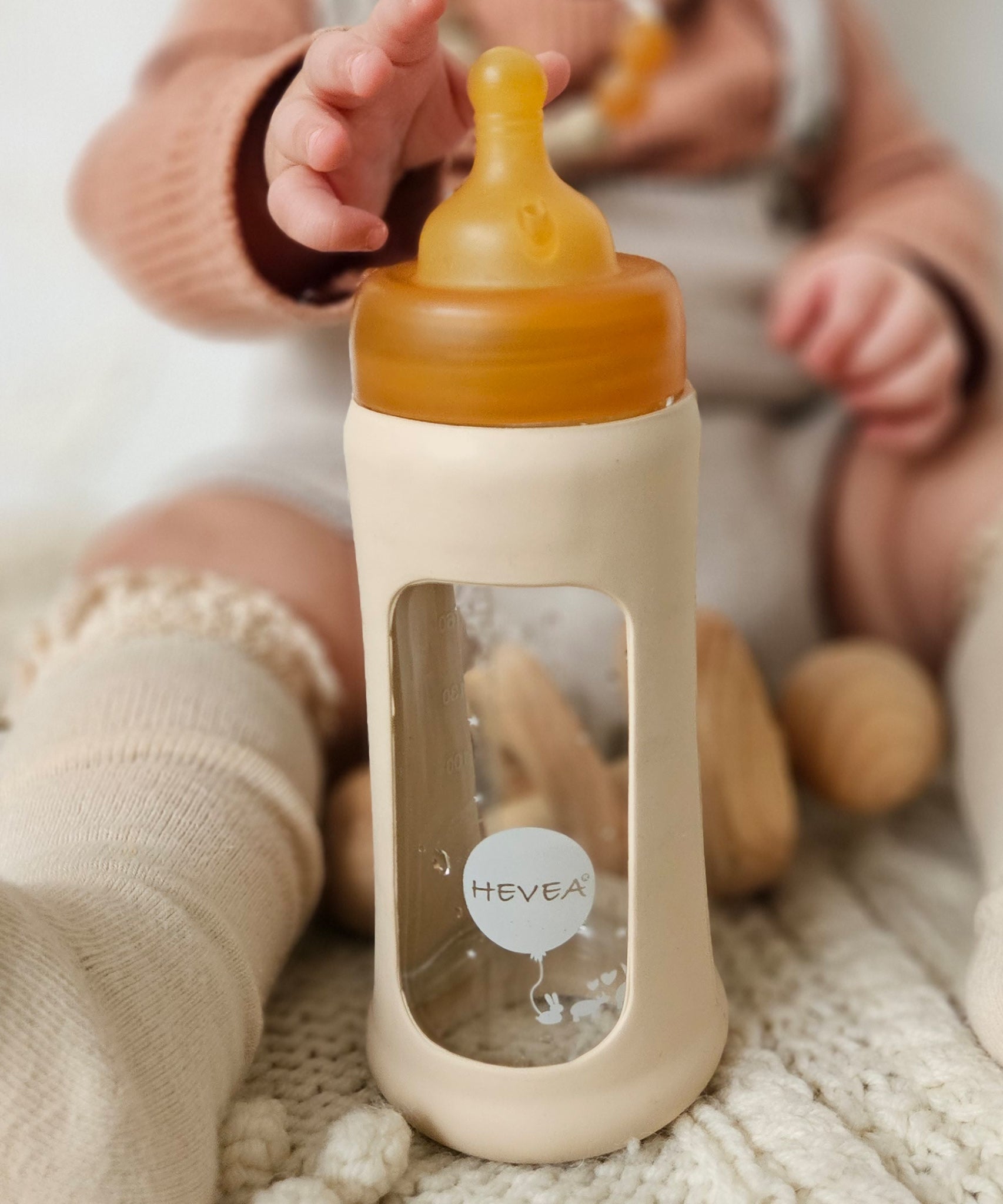 Baby sat on the floor reaching towards the Hevea Glass Bottle Wide Neck With Rubber Sleeve - 250ml in Sand