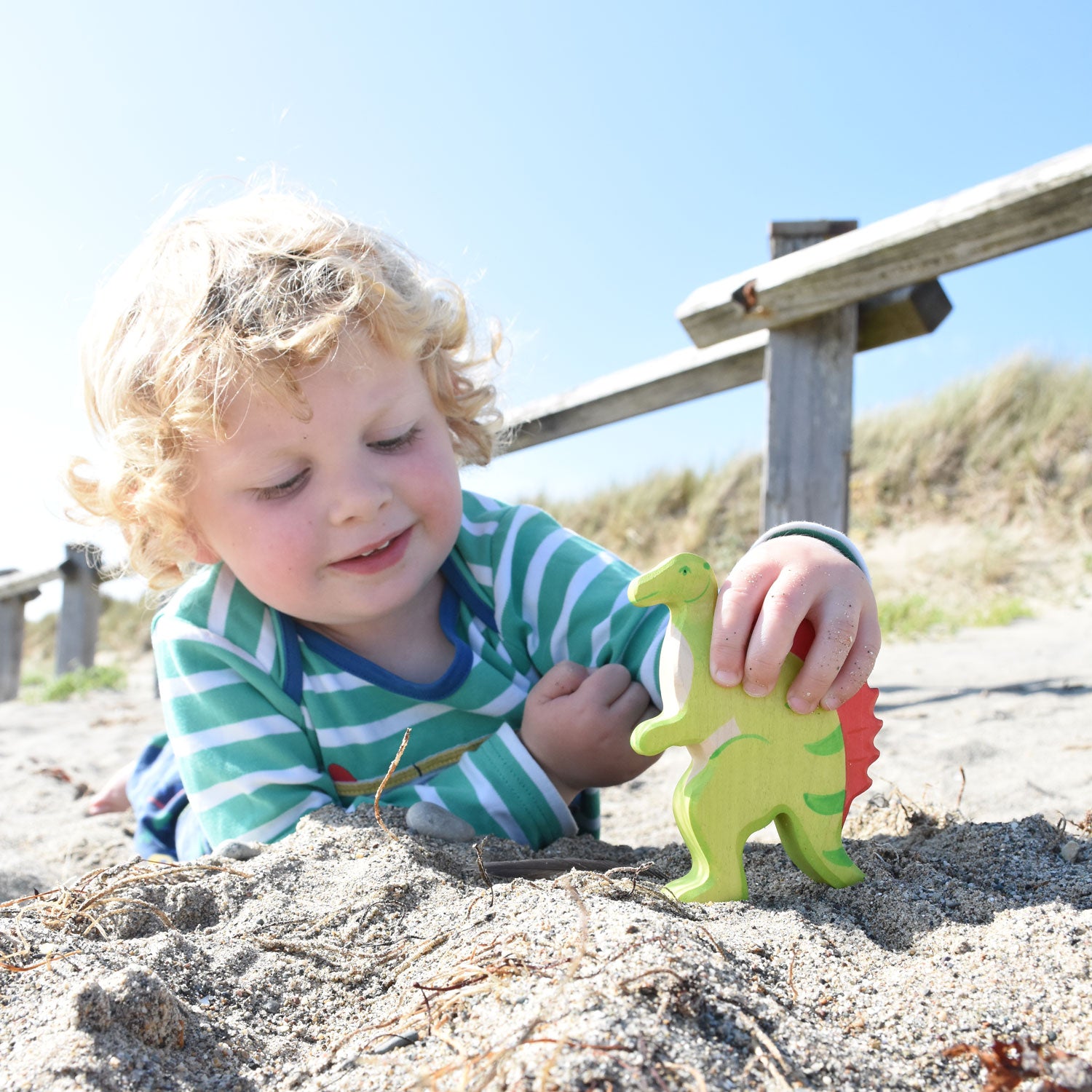 A child lying down on a sandy beach holding onto the Holztiger spinosaurus. The sky is blue and the sunshine is out