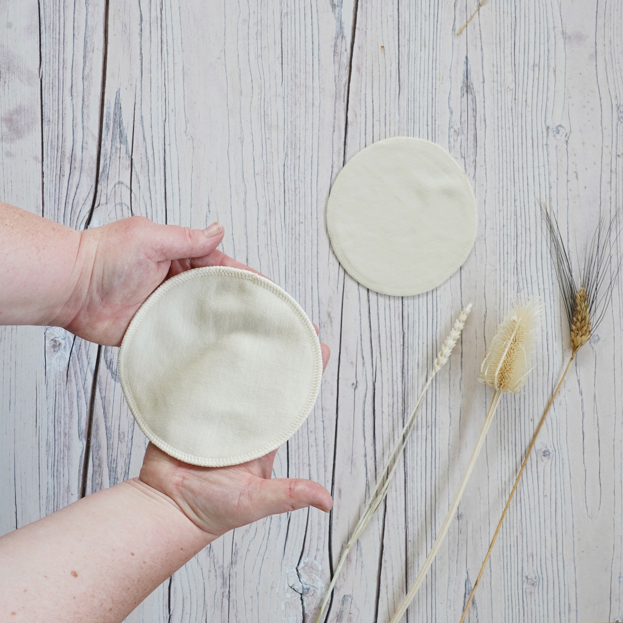 Hands holding one of the Imse Vimse Merino Wool Breast Warmer Pads with the other seen in teh background on a white coloured wooden background 