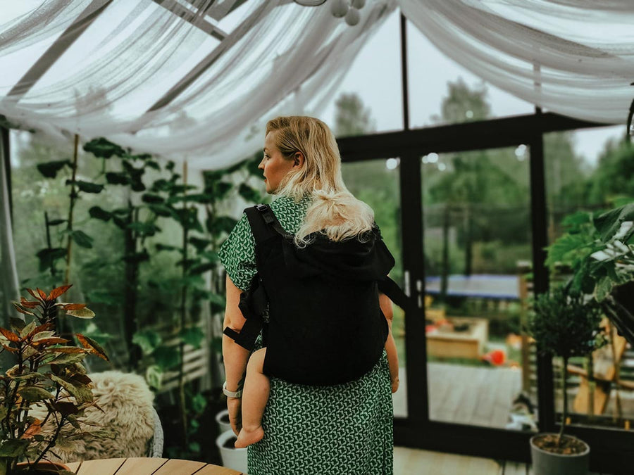 Lady carrying a baby on her back in the black Integra organic linen baby carrier in a garden house