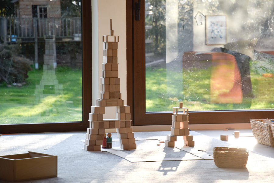 Just Blocks eco-friendly wooden shapes piled into two towers on a grey carpet in front of two large windows