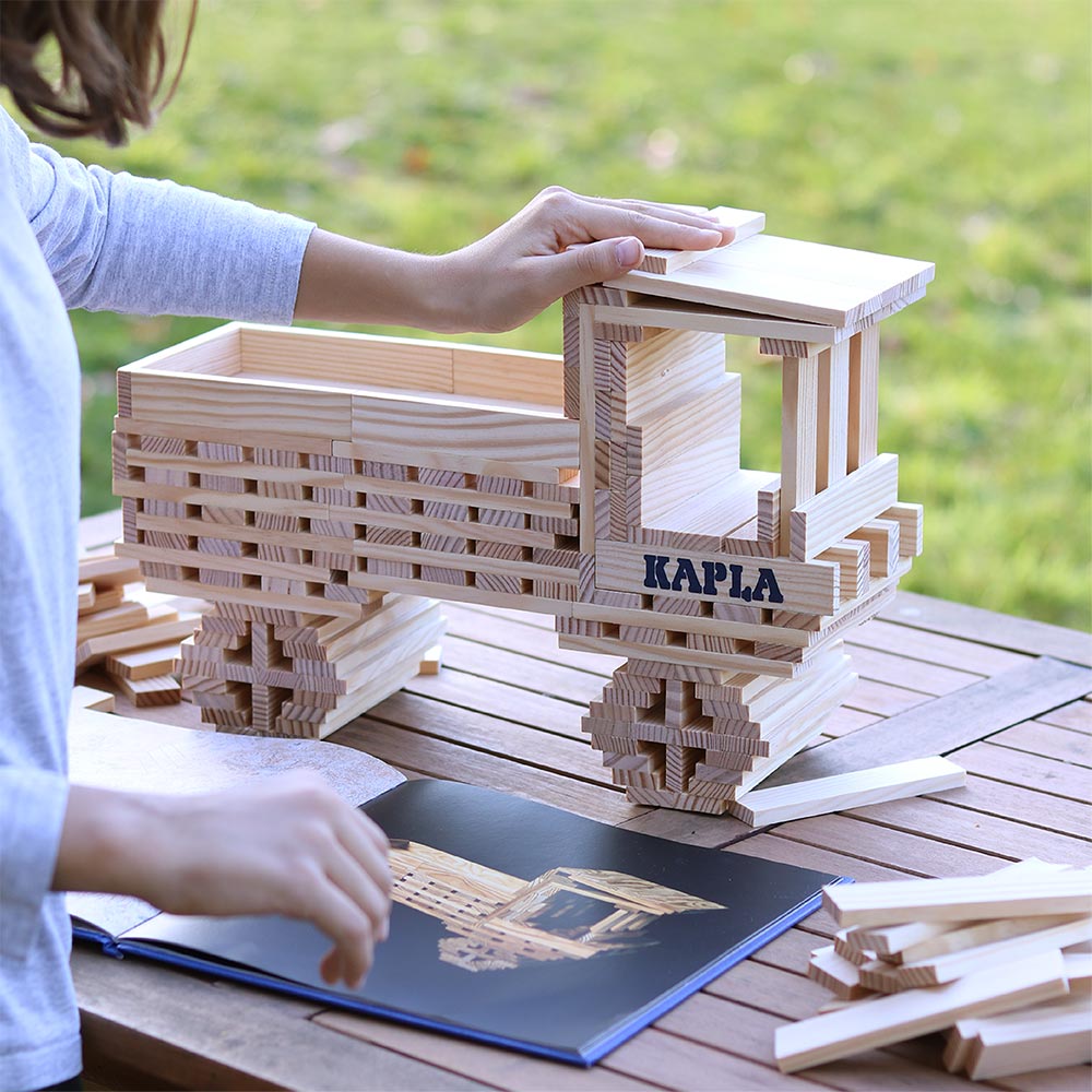 Child placing down a wooden Kapla building block on top of a model truck behind the advanced Kapla art book