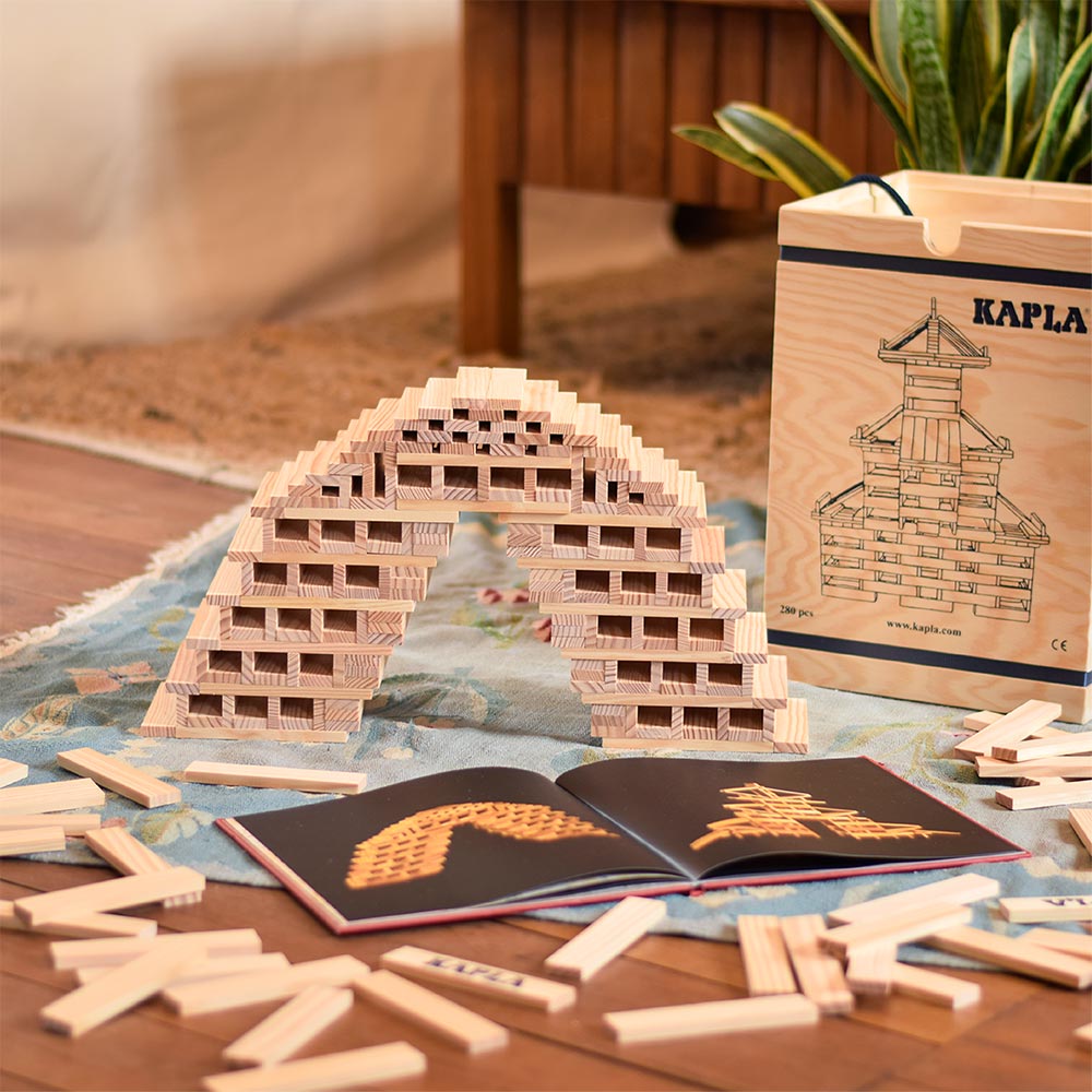 Kapla plastic-free wooden building blocks stacked into a bridge on a wooden floor next to the box