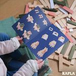 A child holding the leaflet containing build ideas from the KAPLA 200 Box wooden building planks in Spring colours. Wooden blocks can be seen on the floor in the background