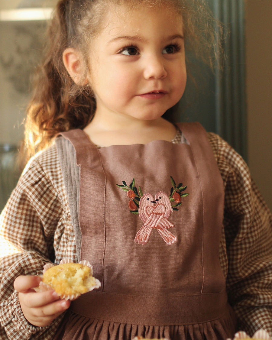 dusky pink pinafore apron with two delicately embroidered pink love birds on the chest panel. Loosely pleated with two pockets on either side of the pinafore and the Avery Row label on the pocket.