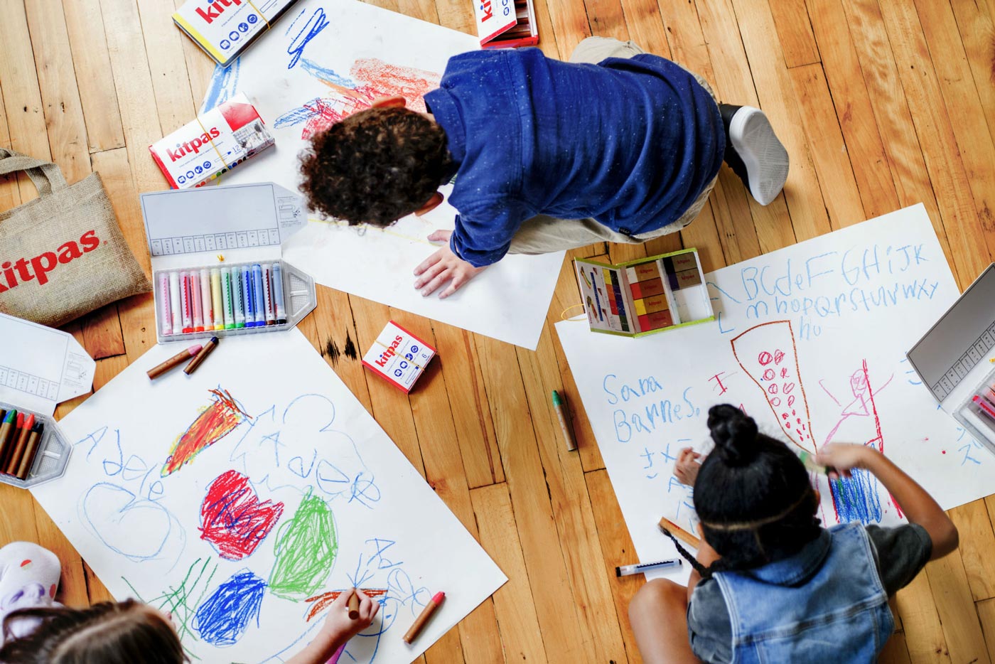 Top down view of children drawing with the Kitpas coloured non-toxic crayons on some large sheets of paper