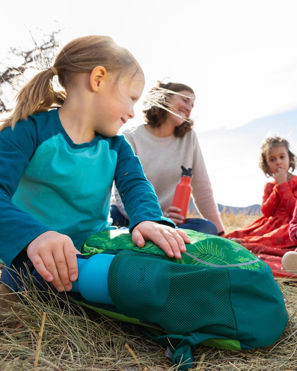 Girl putting a Kid Kanteen 12oz metal bottle into the side pocket of her backpack next to a woman sat on some grass