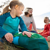 Girl putting a Kid Kanteen 12oz metal bottle into the side pocket of her backpack next to a woman sat on some grass