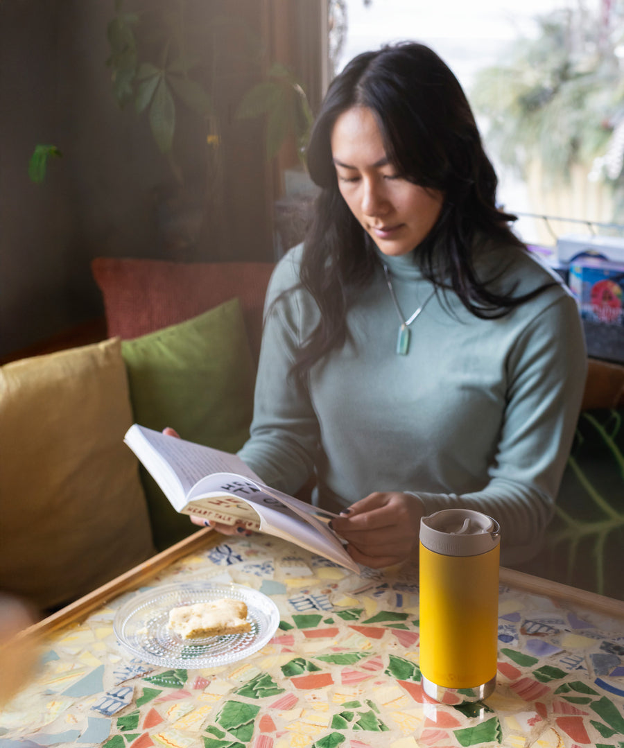 A person sitting at a moisaic covered table reading a book. There is A Klean Kanteen 12oz TKWide Cafe in the sunset yellow colour in front of them.