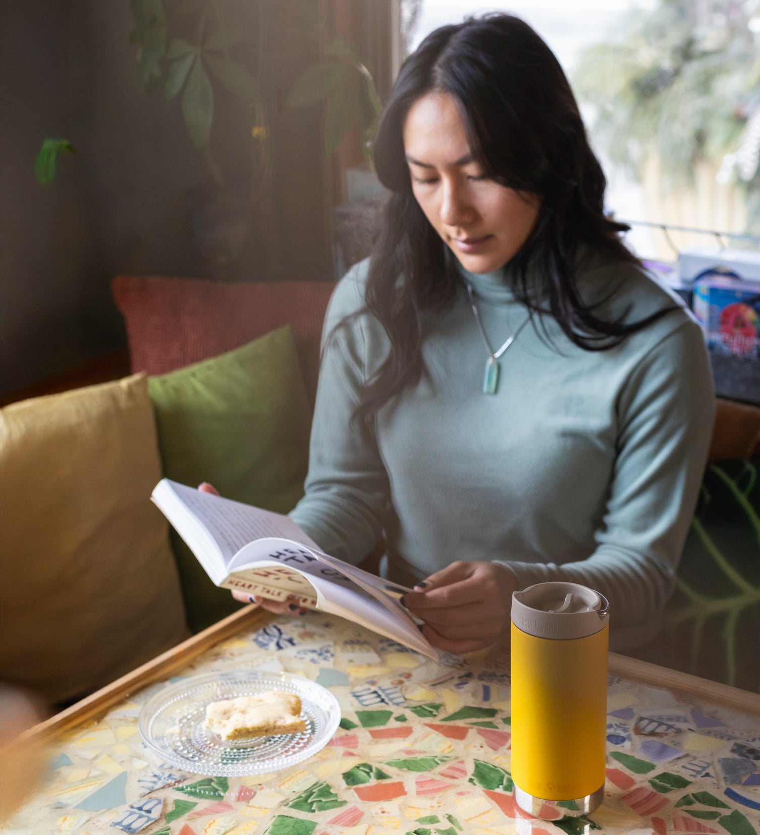 A person sitting at a moisaic covered table reading a book. There is A Klean Kanteen 12oz TKWide Cafe in the sunset yellow colour in front of them.