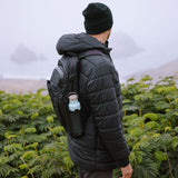 Man stood in front of some green bushes with the Klean Kanteen 20oz insulated classic metal bottle in the side pocket of his backpack