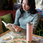 A woman sitting by a table reading a book, there is a peach parfat pink coloured Klean Kanteen 20oz TKWide Cafe on the table n front of her.