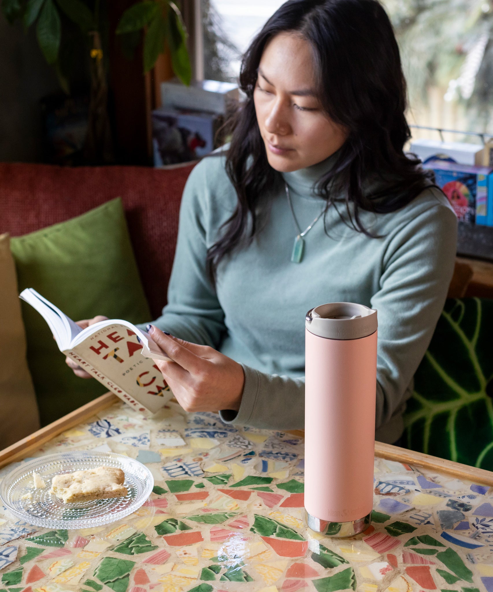 A woman sitting by a table reading a book, there is a peach parfat pink coloured Klean Kanteen 20oz TKWide Cafe on the table n front of her.