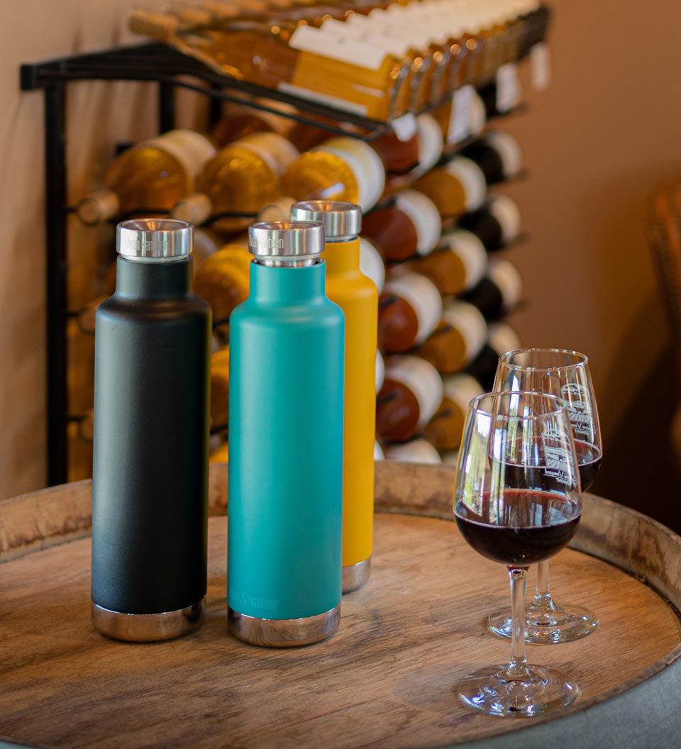 3 klean kanteen 750ml classic insulated bottles with the pour through cap on a wooden barrel next to some glasses of red wine