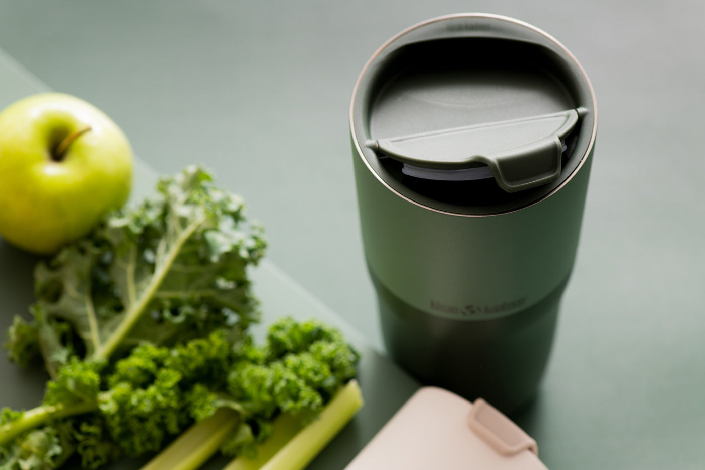 Close up of the flip lid on the Klean Kanteen stainless steel insulated drinks tumbler on a dark green background next to an apple