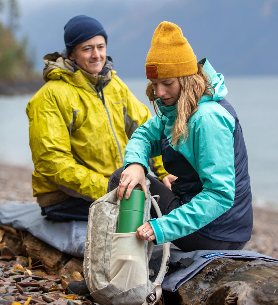 Man and woman sat on a log putting a Klean kanteen 750 ml steel tkpro flask into the side pocket of a rucksack