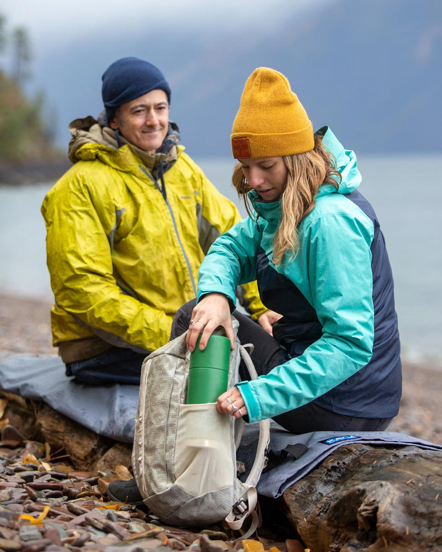 Man and woman sat on a log putting a Klean kanteen 750 ml steel tkpro flask into the side pocket of a rucksack