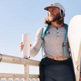 Man leaning on a railing holding a surfboard and a Klean kanteen steel tkpro insulated flask