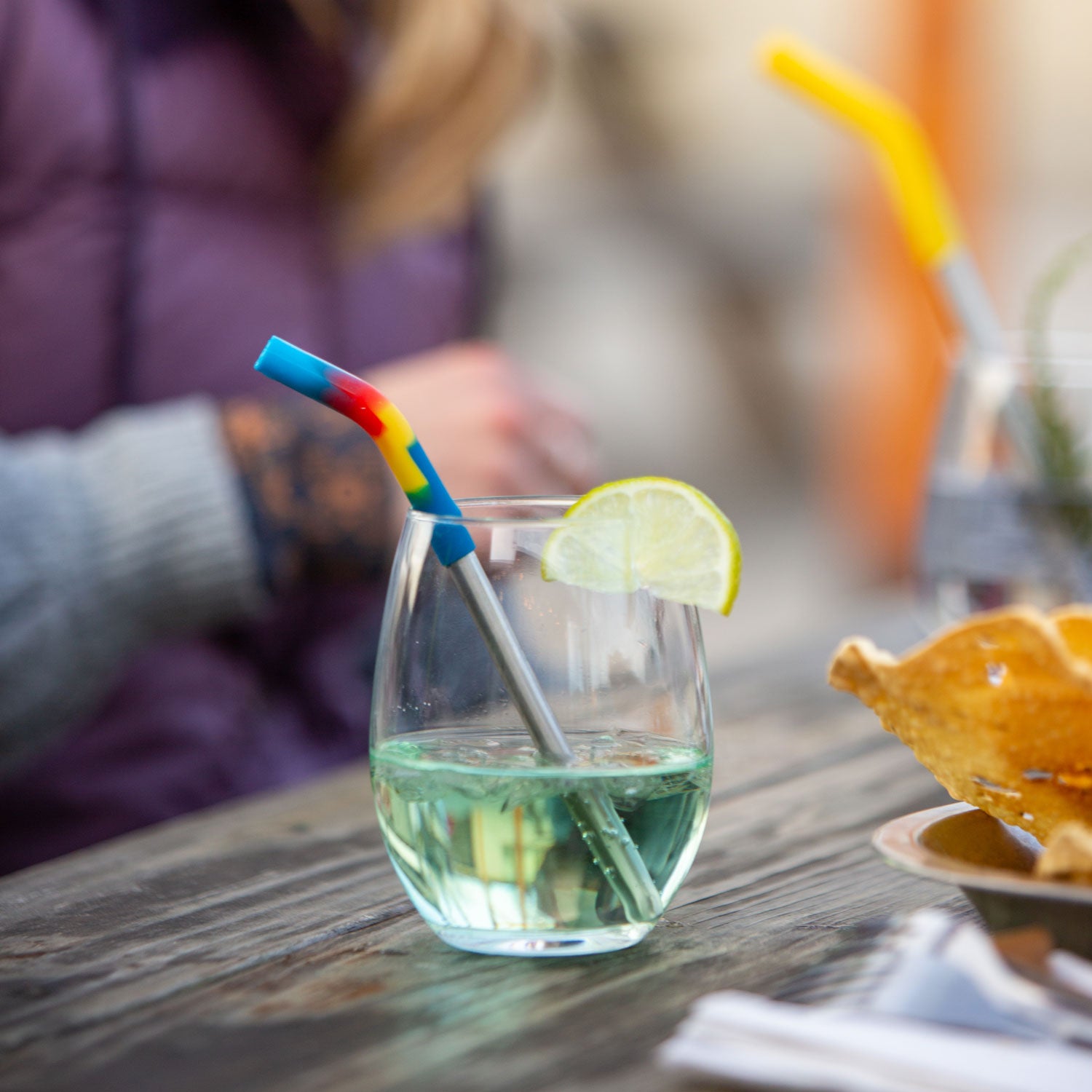 A short glass with a green coloured drink and a lime on the side and a  Klean Kanteen rainbow tipped short straw