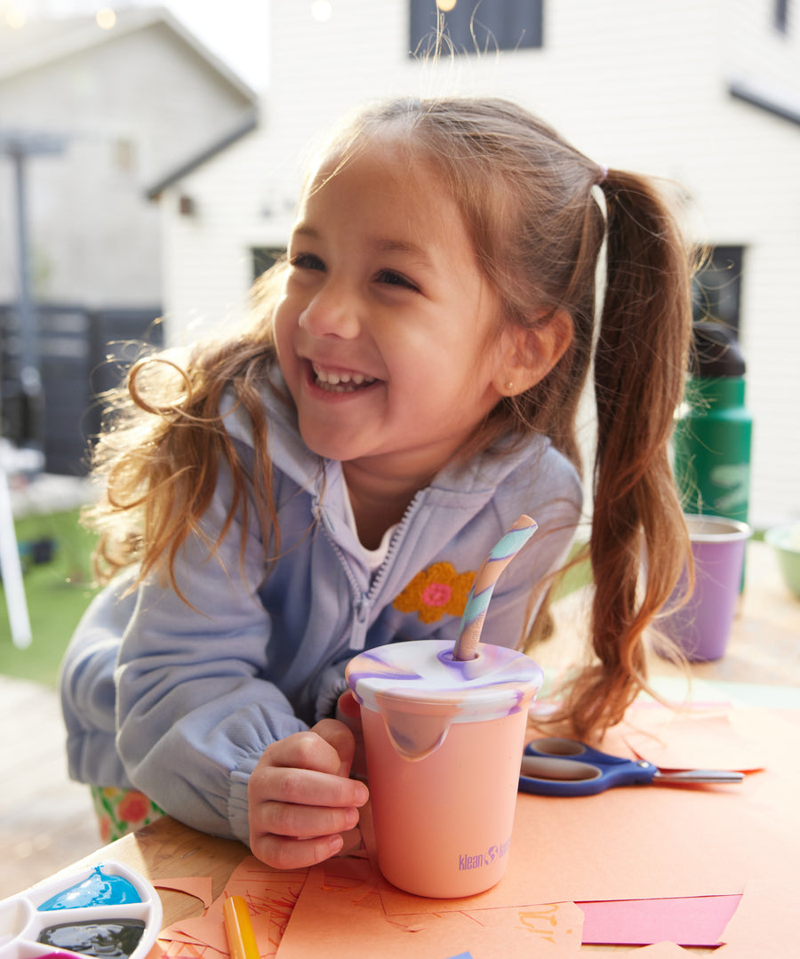 A child smiling with a 10oz Klean Kanteen Steel Kid Cup with Straw Lid in peach parfait pink in front of them. 