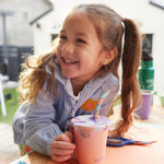 A child smiling with a 10oz Klean Kanteen Steel Kid Cup with Straw Lid in peach parfait pink in front of them. 