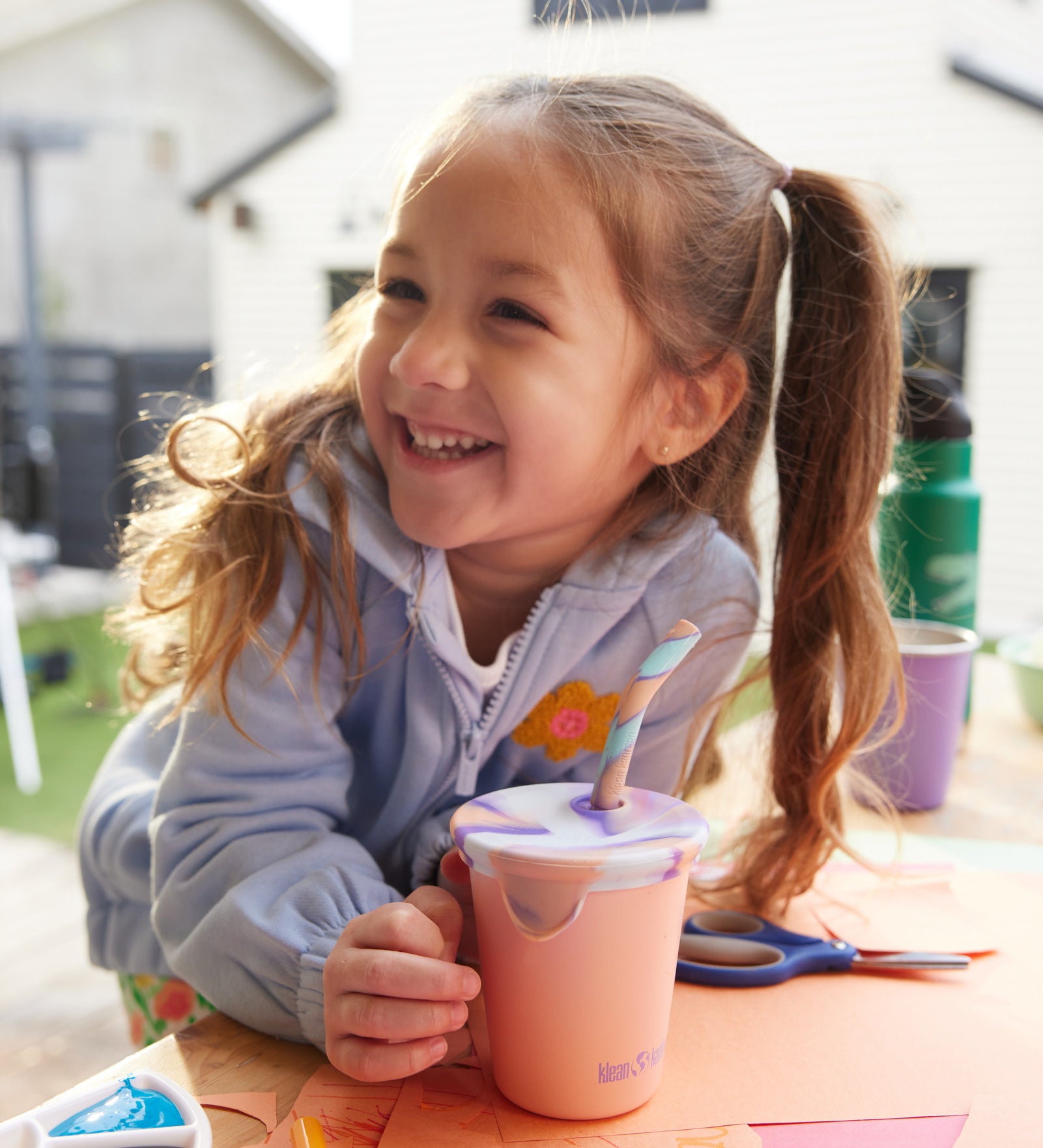 A child smiling with a 10oz Klean Kanteen Steel Kid Cup with Straw Lid in peach parfait pink in front of them. 