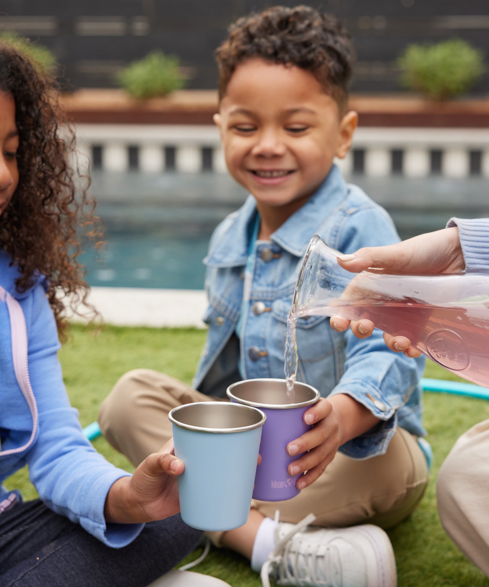 Children holding Klean Kanteen Steel Kid Cups, an adult can be seen pouring raspberry lemonade into the cups. 