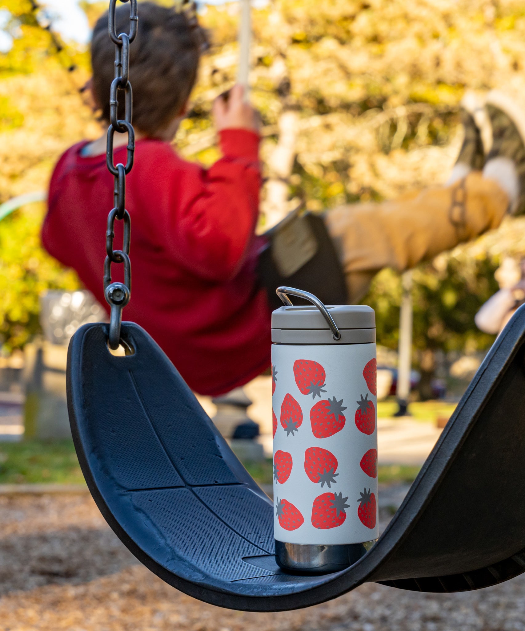 A Klean Kanteen 12oz Tofu Strawberries Print TKWide placed on a swing. A child can be seen swinging on another swing in the background. 