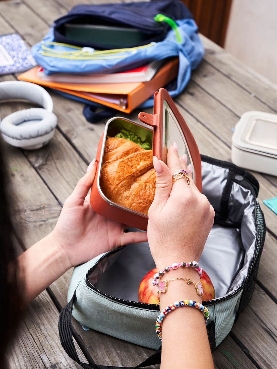 Person opening a Klean Kanteen Rise Stainless Steel Lunch Box with school bags open in the background