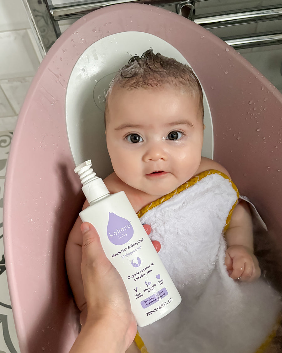 A child sat in a baby bath with a person holding the Kokoso Fragrance Free Hair and Body Wash bottle next to them