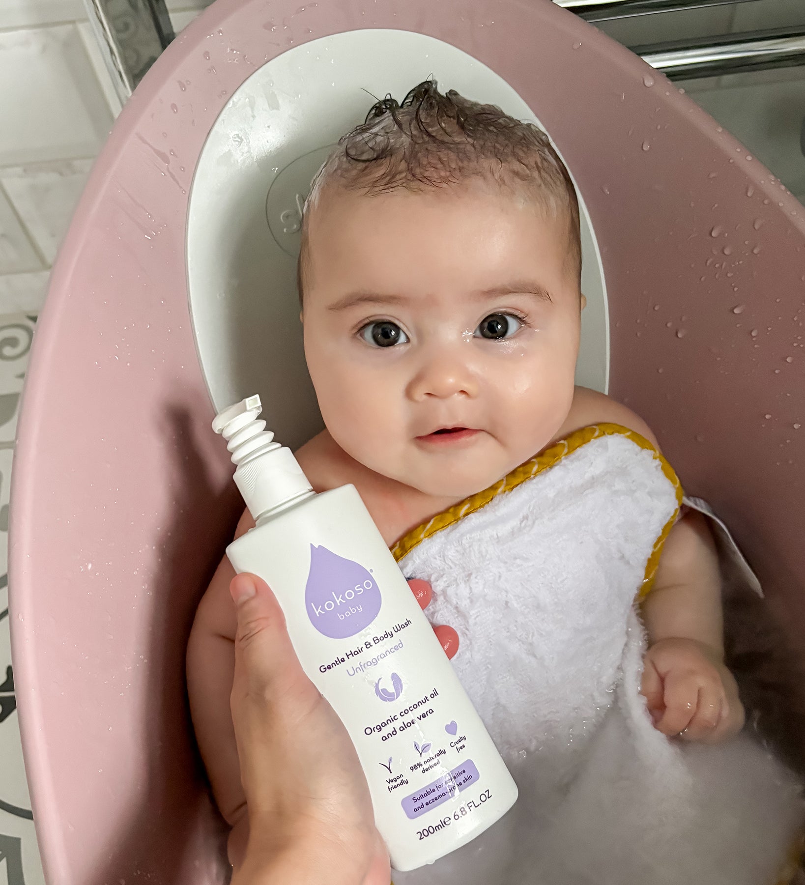 A child sat in a baby bath with a person holding the Kokoso Fragrance Free Hair and Body Wash bottle next to them