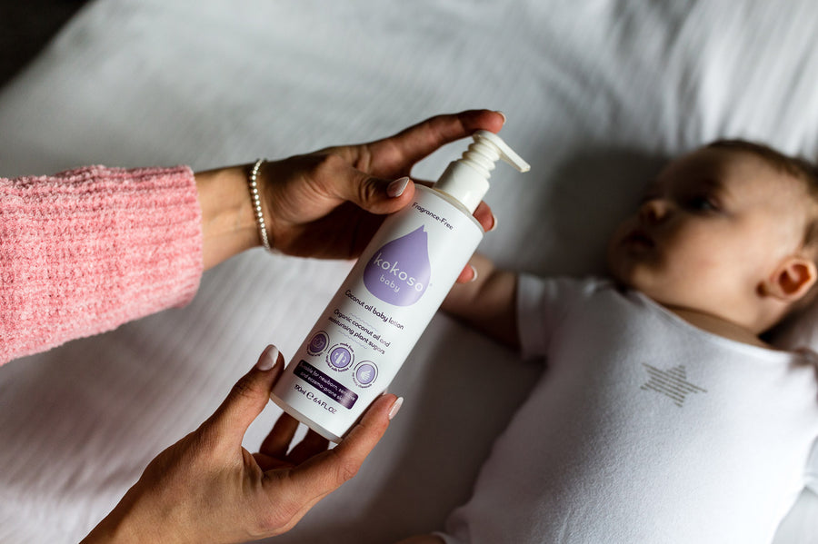 Close up of a woman's hands holding the Kokoso fragrance-free coconut oil lotion above a baby laying on a white bed