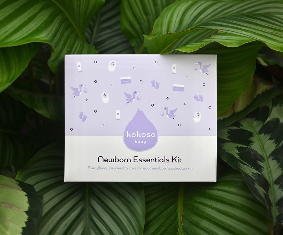 The reverse of the Kokoso Newborn Essentials Organic Baby Gift Set in its purple and white gift box sitting on a backdrop of large Green leaves. The words "Pure and gentle skincare for brand new babies" stand out.