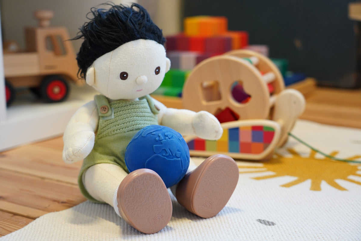 Close up of an Olli Ella Dinkum Doll toy sat on a white carpet with a Lanco natural rubber globe ball in its lap