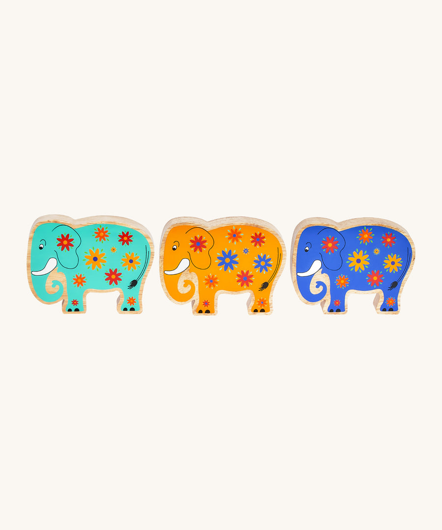 A green, yellow and blue wooden Lanka Kade elephants standing next to each other on a cream background