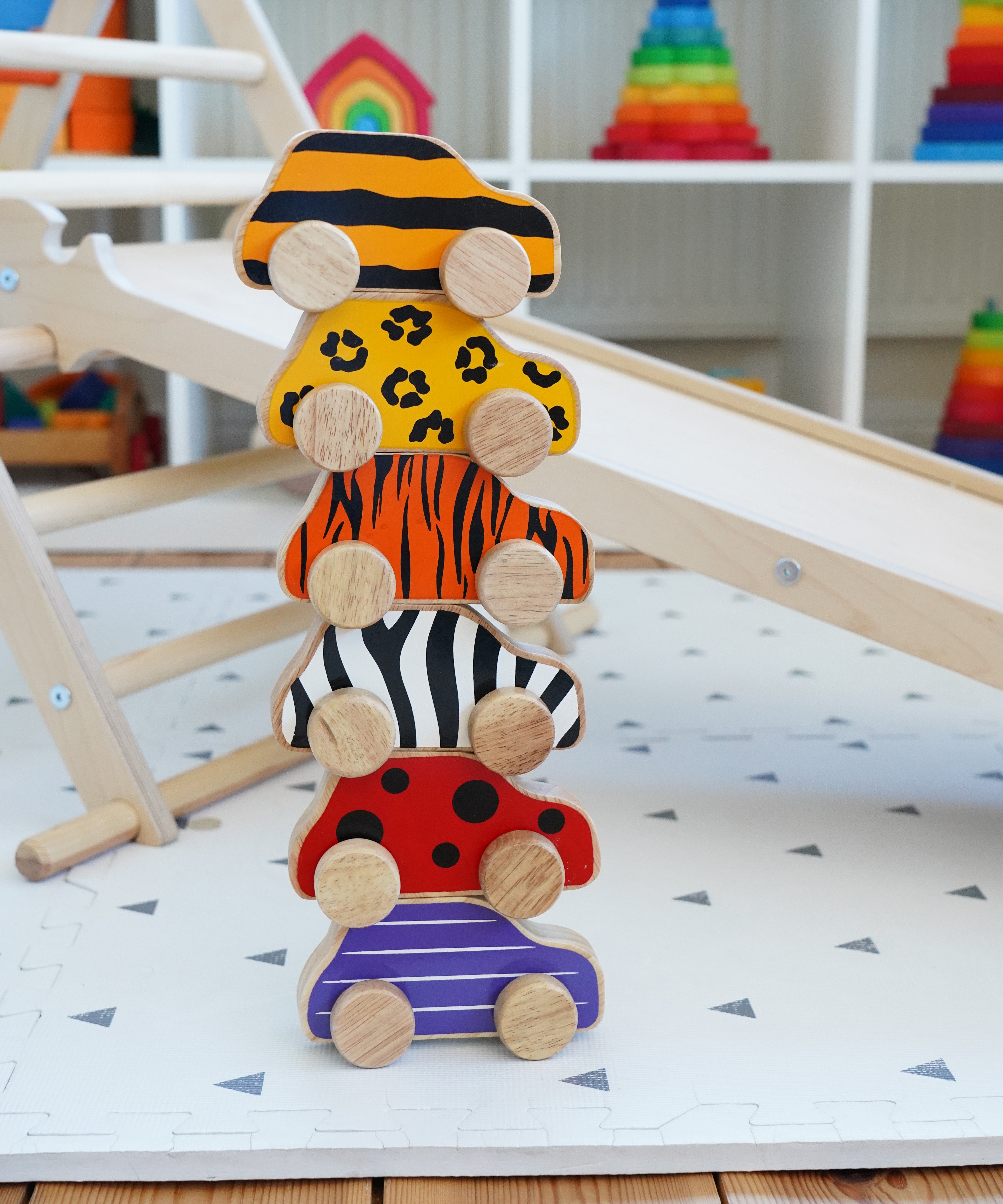 Lanka Kade wooden animal printed cars stacked on a playmat next to a Triclimb wooden slide and wooden climbing frame. Features a bumblebee, leopard, tiger, zebra, ladybird and beetle printed wooden car.