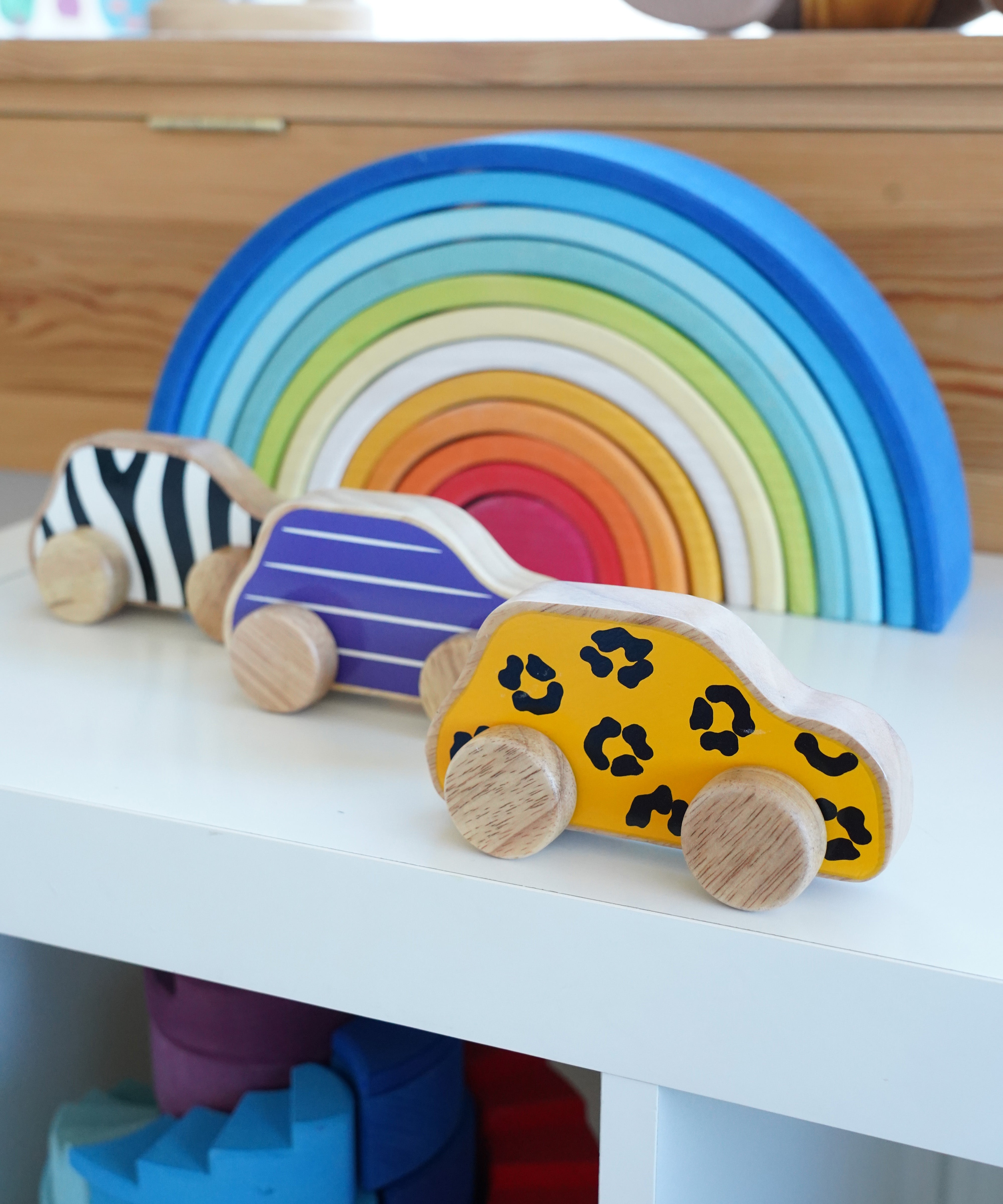 Lanka Kade wooden animal printed cars next to a Grimms rainbow on a shelf. Features a leopard, snail and zebra printed wooden car. 