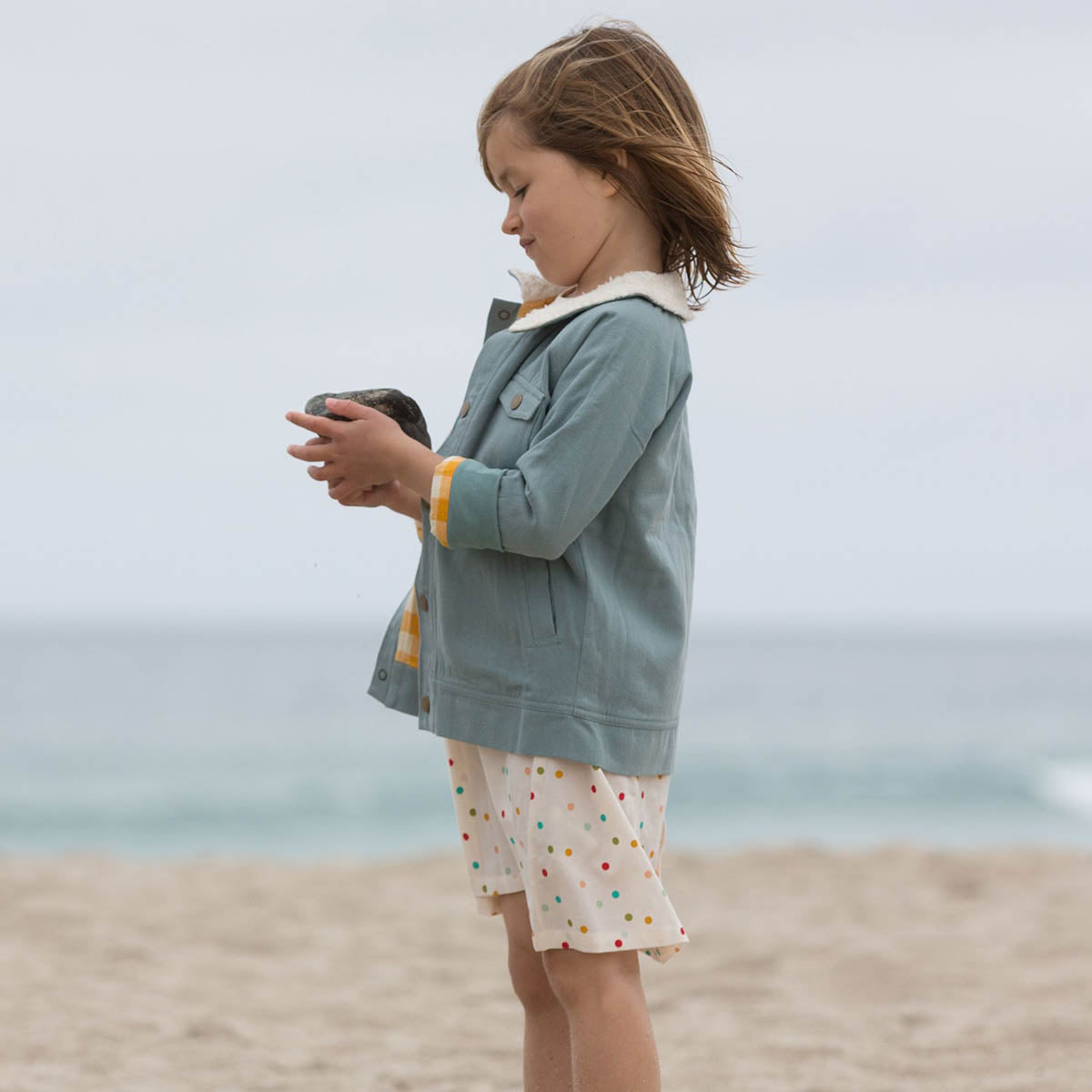 Young girl stood on a beach wearing a spotted dress and a blue Little Green Radicals sherpa collar jacket