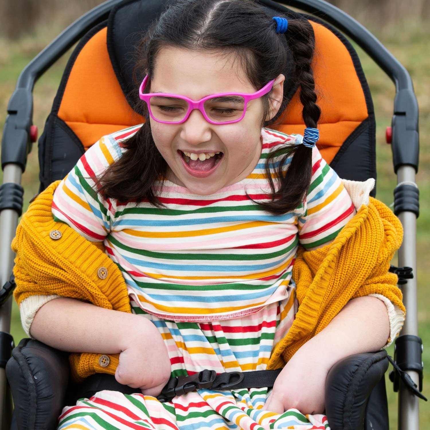 Child wearing the Little Green Radicals Adaptive Rainbow Stripe Easy Peasy Dress with a yellow knitted cardigan 