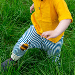 A close up of a child's legs wearing the Little Green Radicals Bees Knees Patch Striped blue and white Joggers. The child is also wearing a yellow gold coloured short sleeved t-shirt whilst running through long green grass.