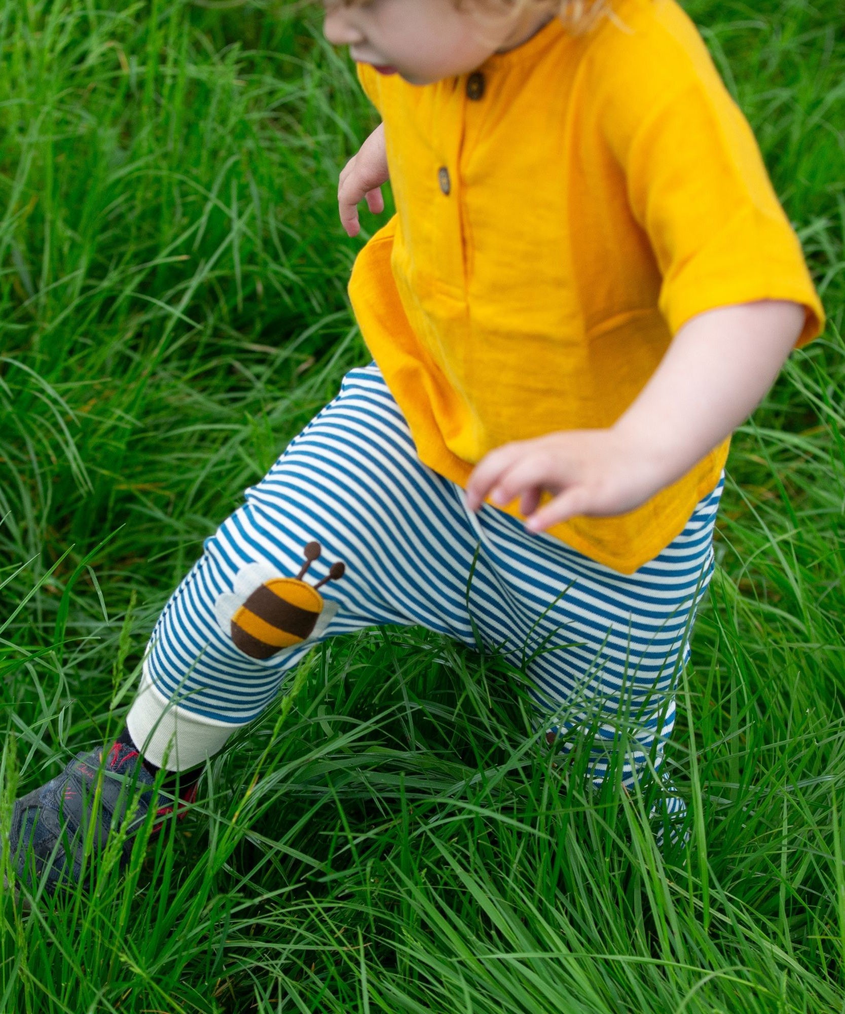 A close up of a child's legs wearing the Little Green Radicals Bees Knees Patch Striped blue and white Joggers. The child is also wearing a yellow gold coloured short sleeved t-shirt whilst running through long green grass.