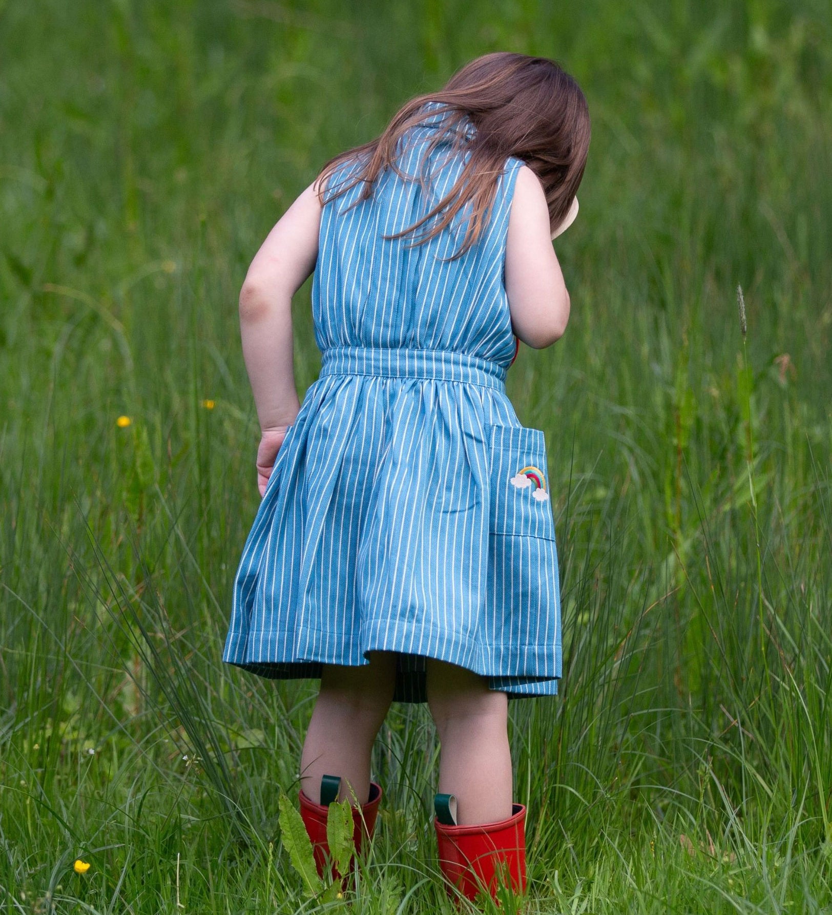 A child wearing the Little Green Radicals Child's Organic Cotton Horizon Blue Striped Pinafore Button Dress with red wellington boots. 