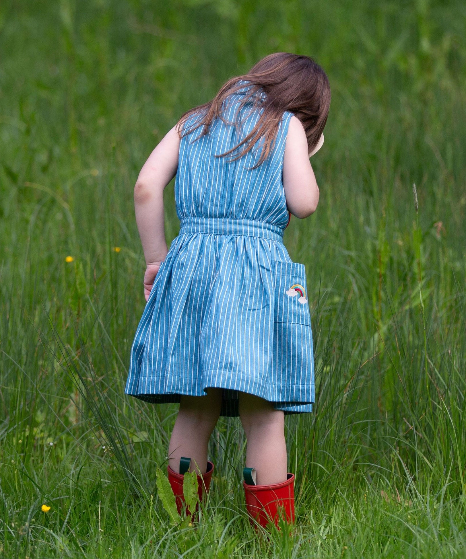 A child wearing the Little Green Radicals Child's Organic Cotton Horizon Blue Striped Pinafore Button Dress with red wellington boots. 