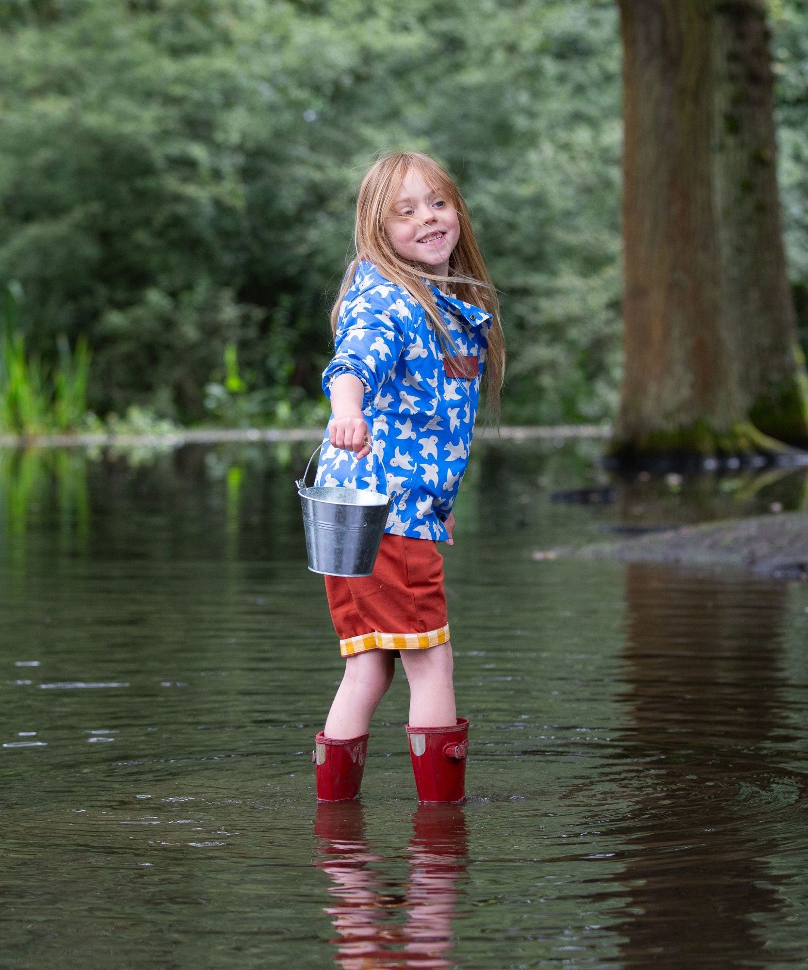 A side view of a child wearing the Little Green Radicals Diving And Splashing Blue Recycled Waterproof Kids Packable Anorak