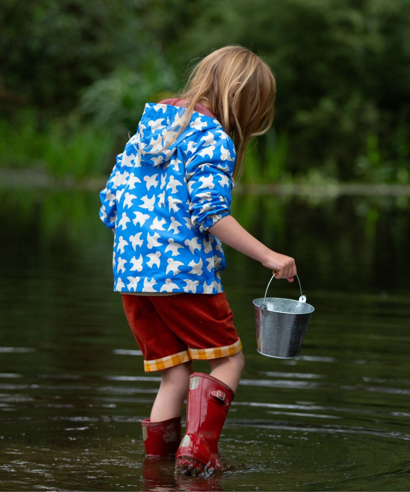 A child wearing the Little Green Radicals Diving And Splashing Blue Recycled Waterproof Kids Packable Anorak, facing away from the camera showing the back of the jacket.