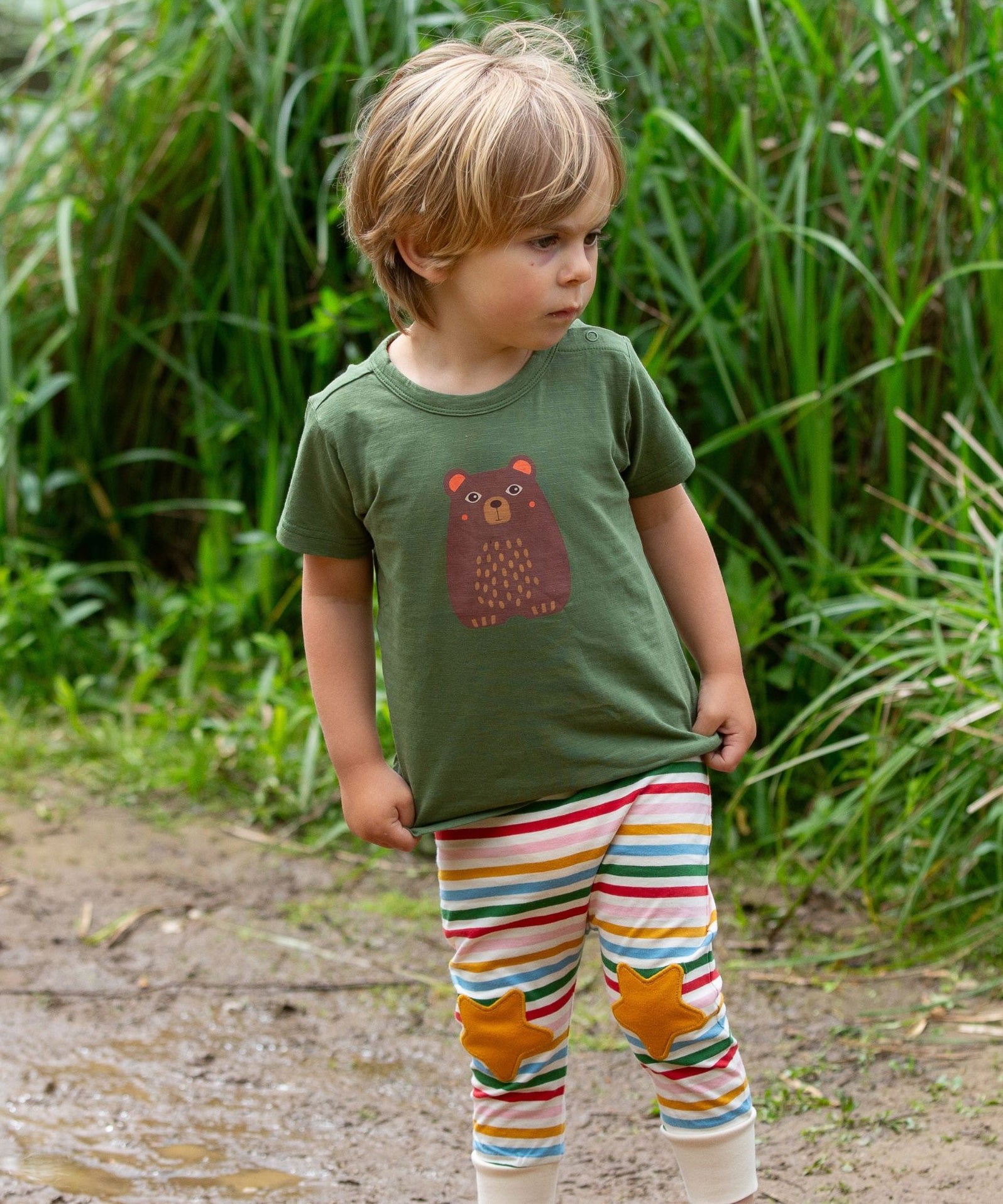 A child wearing the Little Green Radicals Kids Organic Cotton Green Bear Short Sleeve T-Shirt with rainbow striped star knee patch joggers.