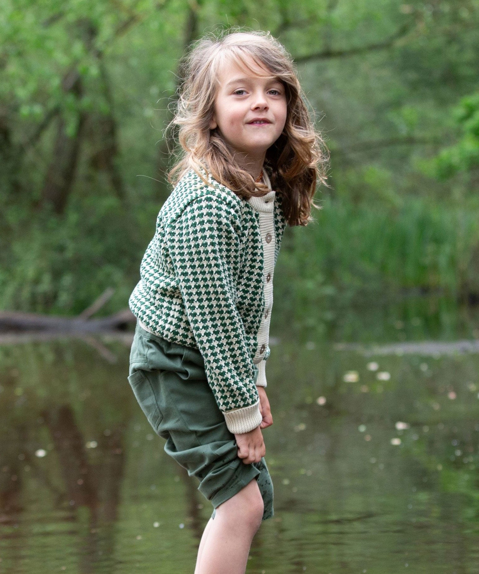 A child wearing the Little Green Radicals From One To Another Olive Green Striped Knitted Child's Organic Cotton Cardigan with green coloured shorts.